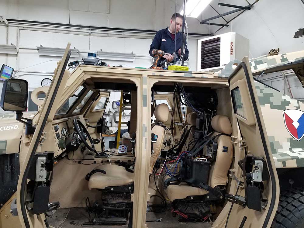 An engineer installing and testing a liquid and air cleaning system on a Lidar sensor mounted on a military vehicle, ensuring optimal performance in rugged environments.